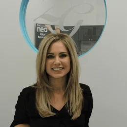 A woman with long blonde hair wearing a black top stands and smiles in front of a circular glass window with the letters "ZG" and "EMSCULPT neo" visible.