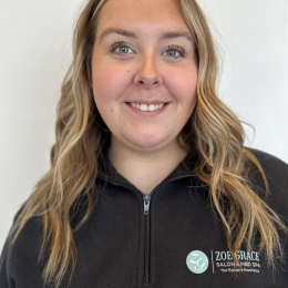 A woman with long wavy blonde hair smiles at the camera, wearing a black zip-up top with a Zoe Grace Salon & Med Spa logo on it, standing against a plain white background.
