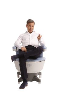 A man in a white shirt and dark pants sits with legs crossed in a modern medical chair, pointing forward, against a plain white background.