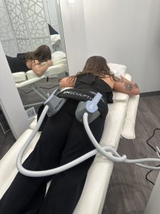 A person lies face down on a treatment bed with EMSCULPT device attachments on their lower back in a clinical setting.