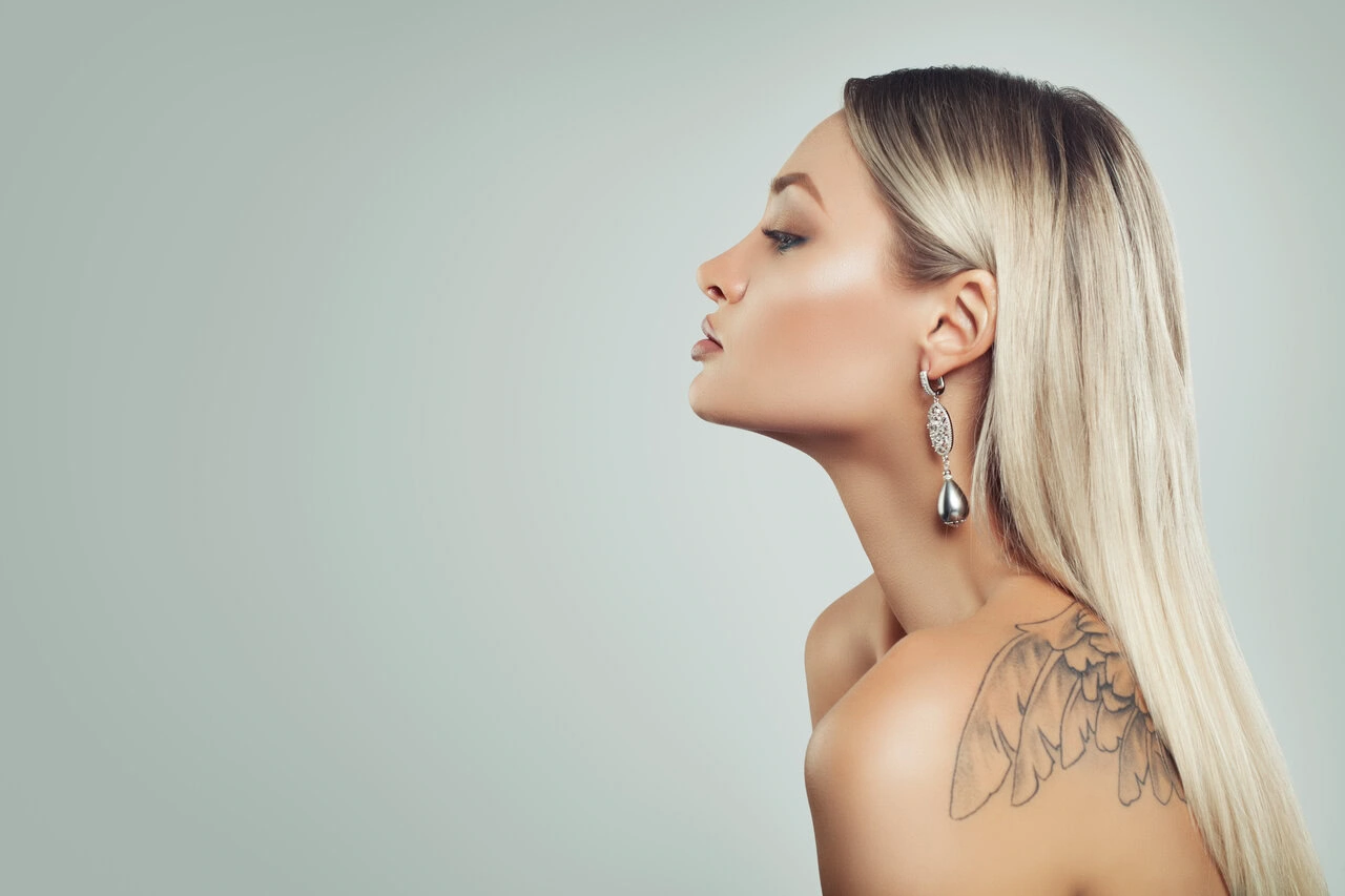 Woman in profile with straight blonde hair, wearing a dangling earring and showing a wing tattoo on her shoulder, against a neutral background.