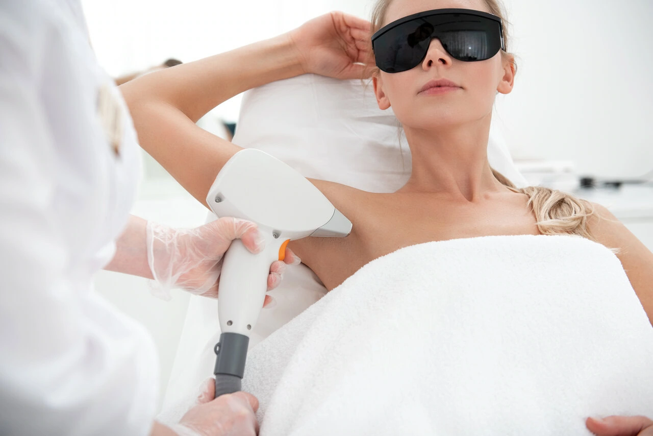 A woman wearing protective goggles receives a laser hair removal treatment on her armpit from a technician in a clinical setting.