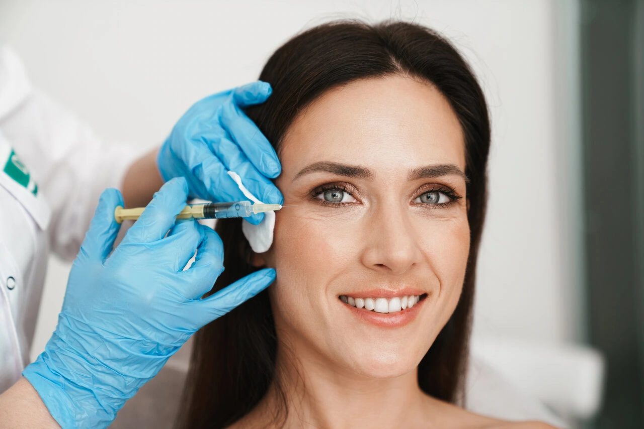 A woman receives a cosmetic injection near her eye from a person wearing blue gloves.
