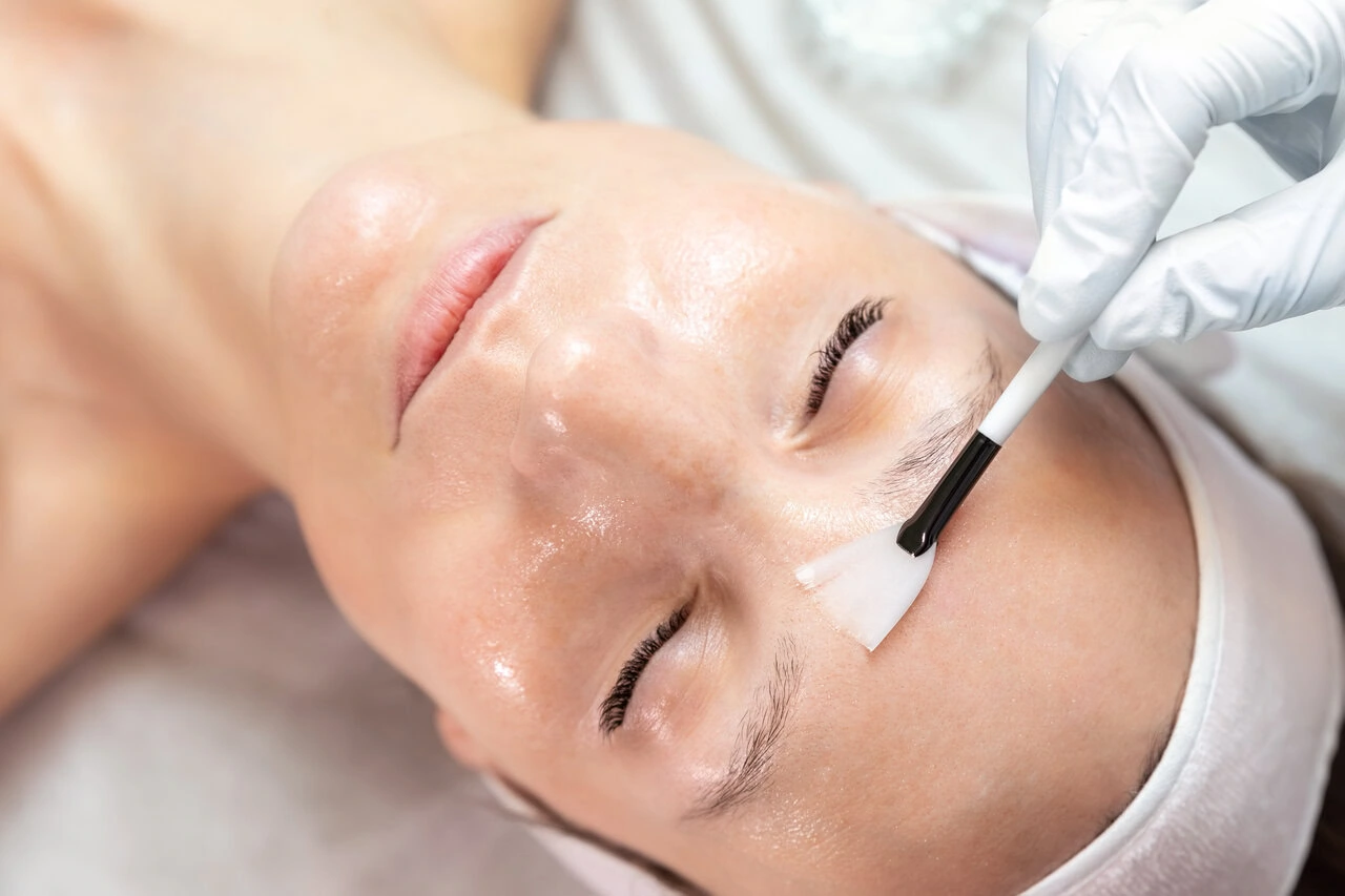 A person lying down with eyes closed receives a facial treatment as a gloved hand applies a product to their forehead with a small brush.