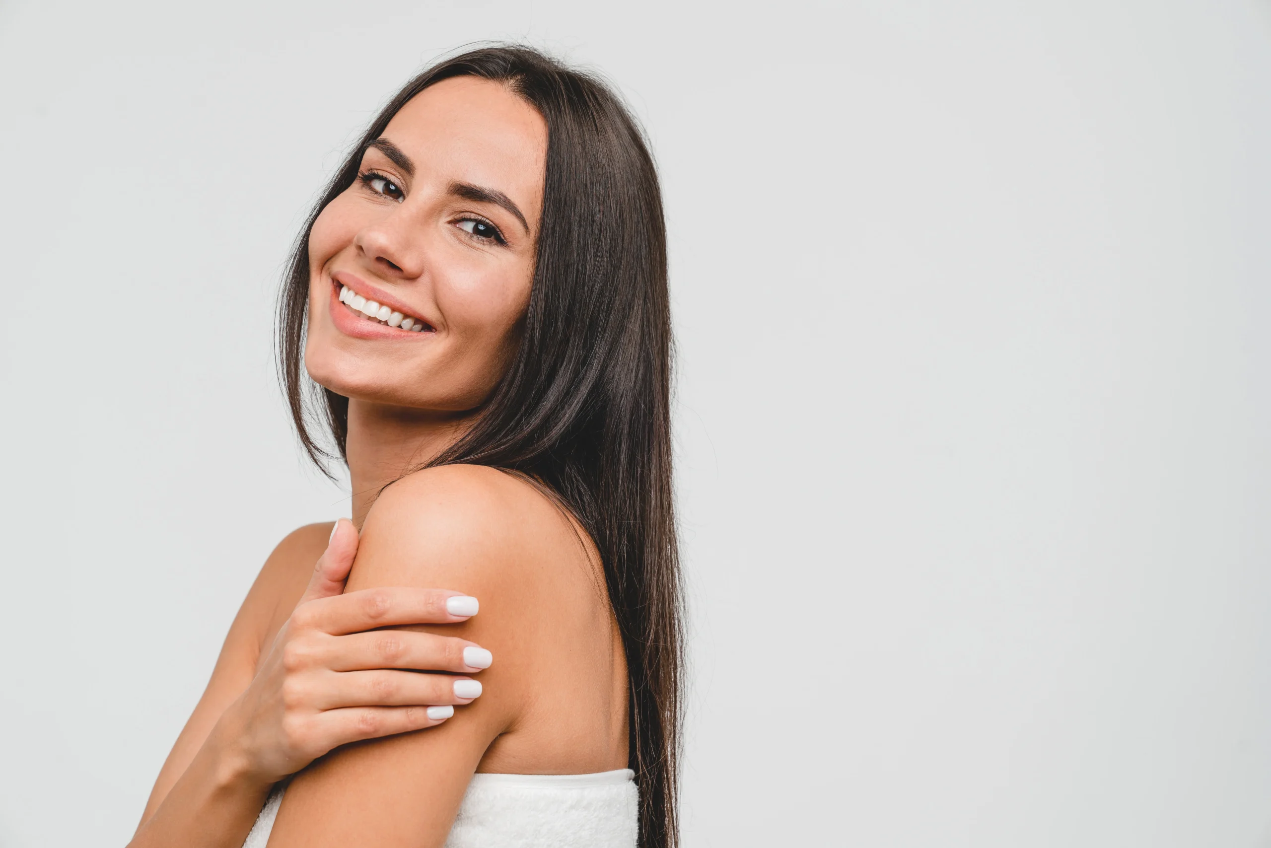 Woman with long dark hair smiling, facing the camera with her head turned sideways, wearing a white top, and touching her shoulder against a plain light background.