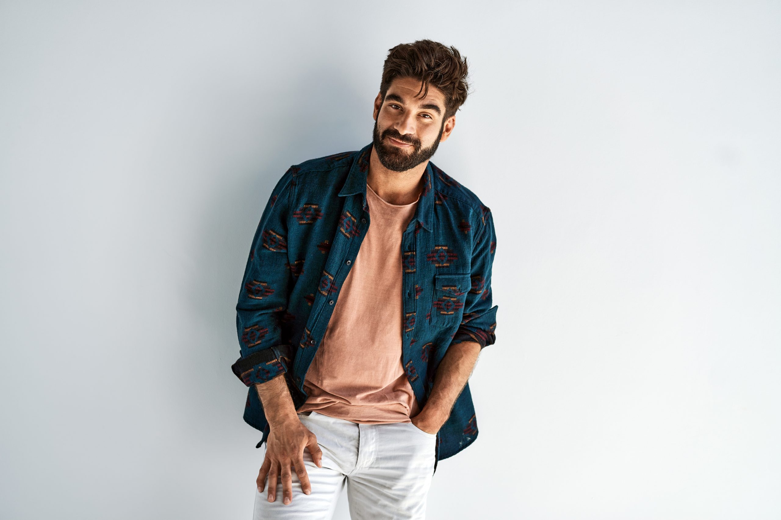 Man with dark hair and beard wearing a blue patterned shirt over a peach t-shirt and white pants, standing against a plain light background.