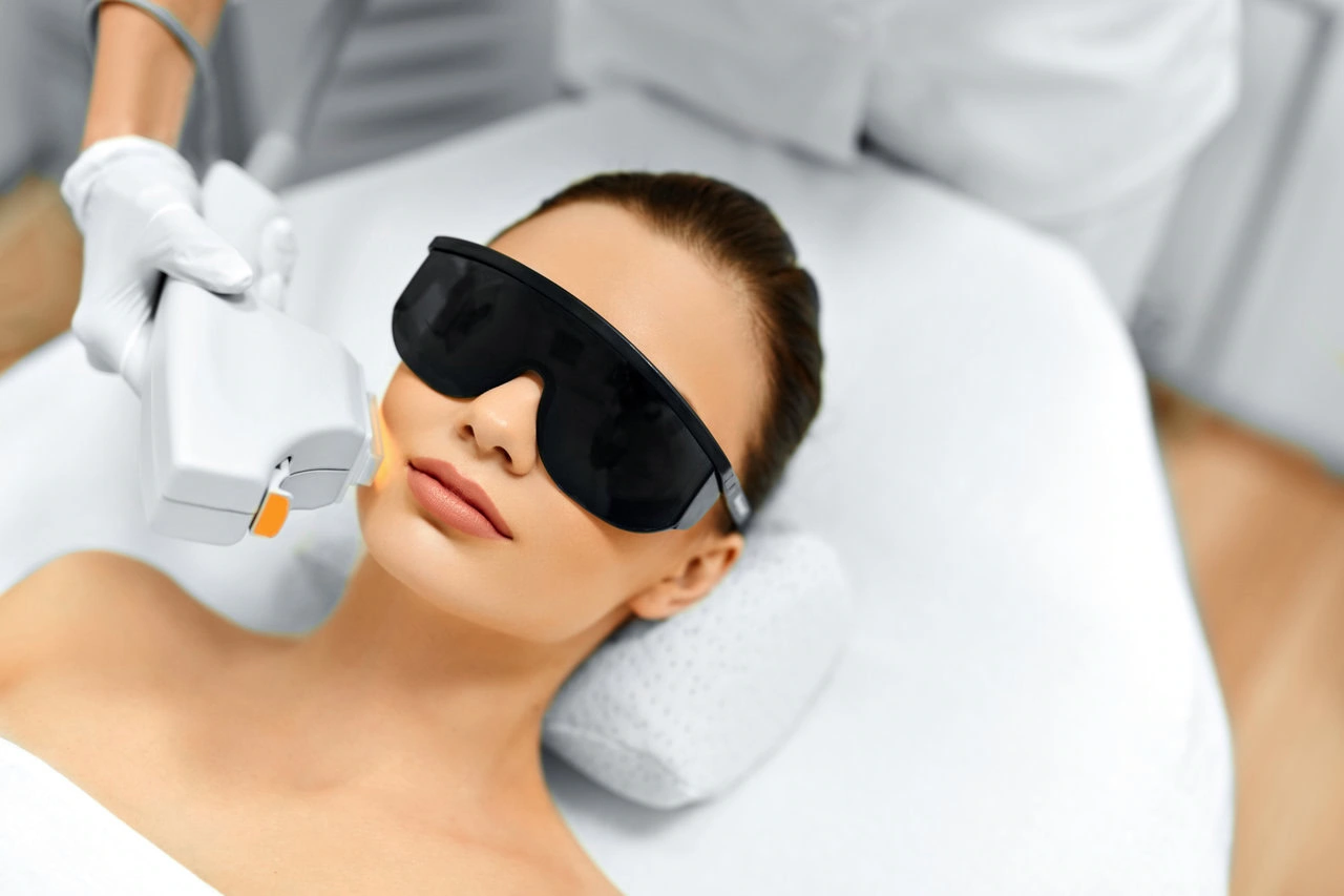 Person lying on a treatment bed wearing protective goggles while receiving a laser facial procedure from a gloved technician.
