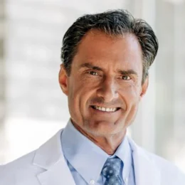 Man wearing a white lab coat, light blue shirt, and blue patterned tie, standing indoors and smiling at the camera.