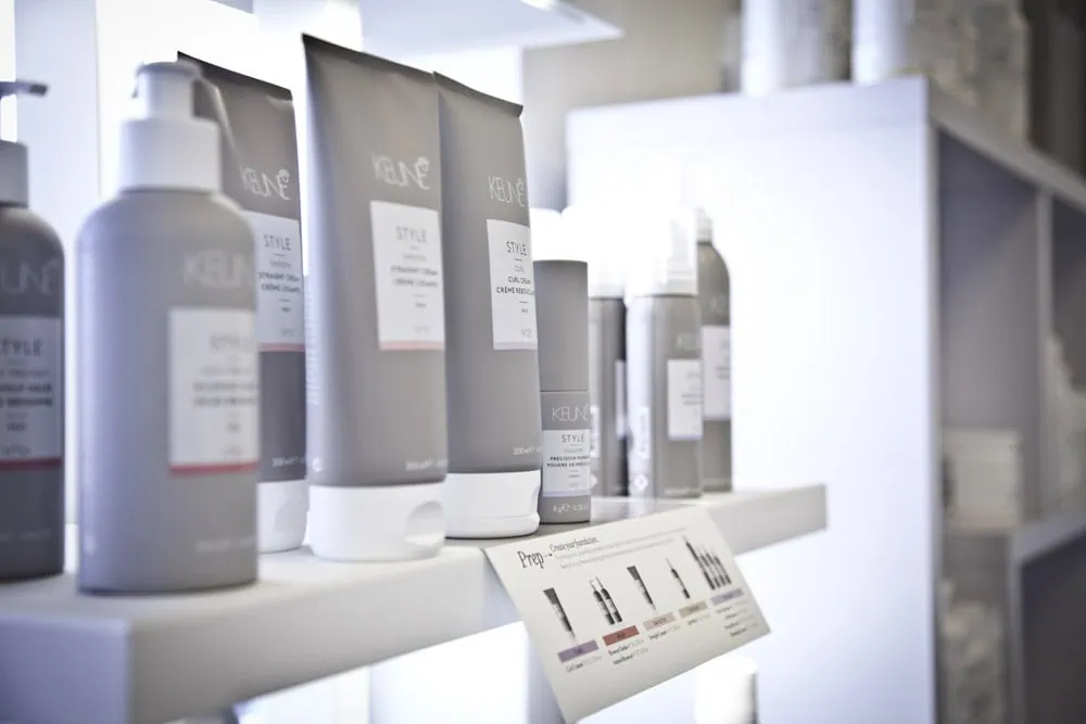 Several grey bottles and tubes of hair care products are displayed on white shelves in a bright, modern retail setting.
