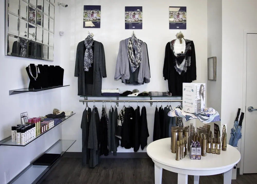 A boutique displays clothing, scarves, jewelry, and beauty products on shelves, racks, and a round table in a clean, modern setting.