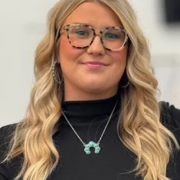 A woman with long blonde hair, wearing glasses, a black top, turquoise earrings, and a matching necklace, stands in front of a blurred light background.