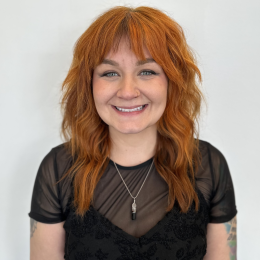 A woman with wavy, shoulder-length auburn hair smiles at the camera. She is wearing a black lace top, a sheer black shirt, and a pendant necklace, standing against a plain white background.