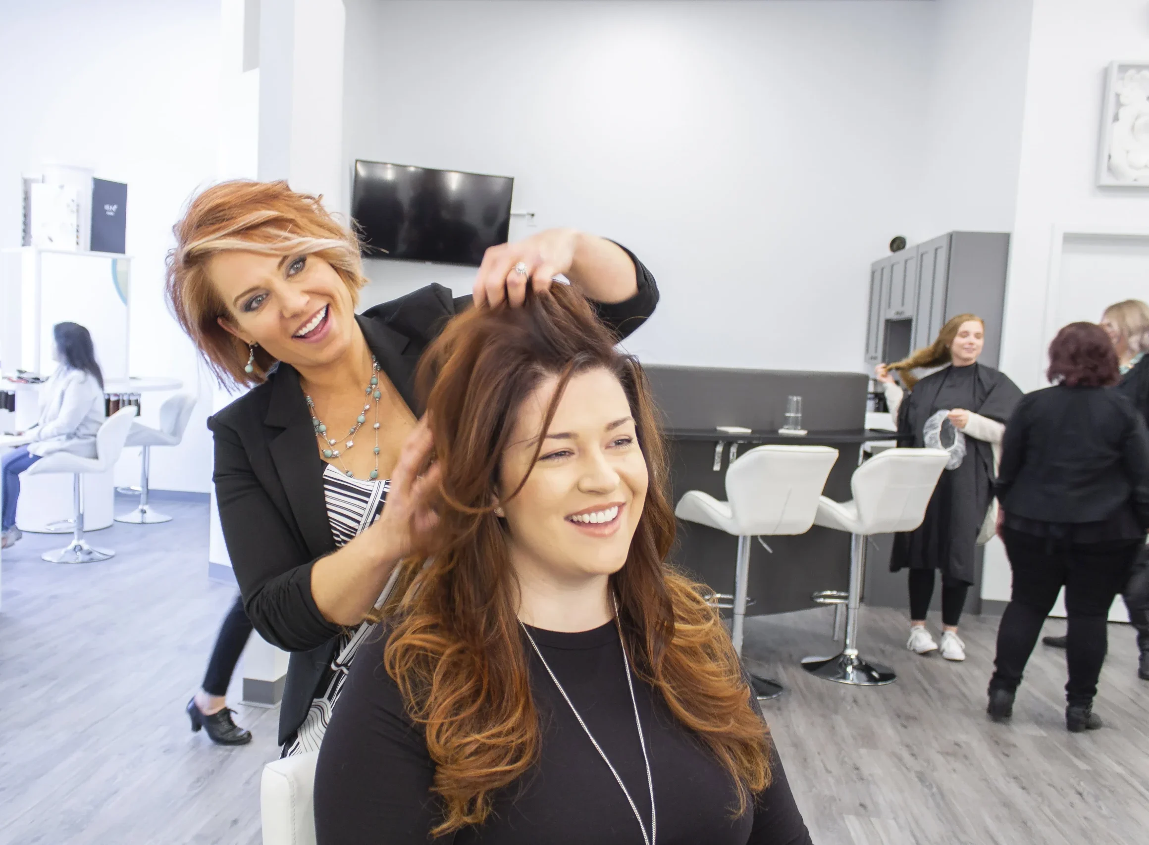 A hairstylist styles a smiling woman's hair in a modern, bright salon with other people working in the background.