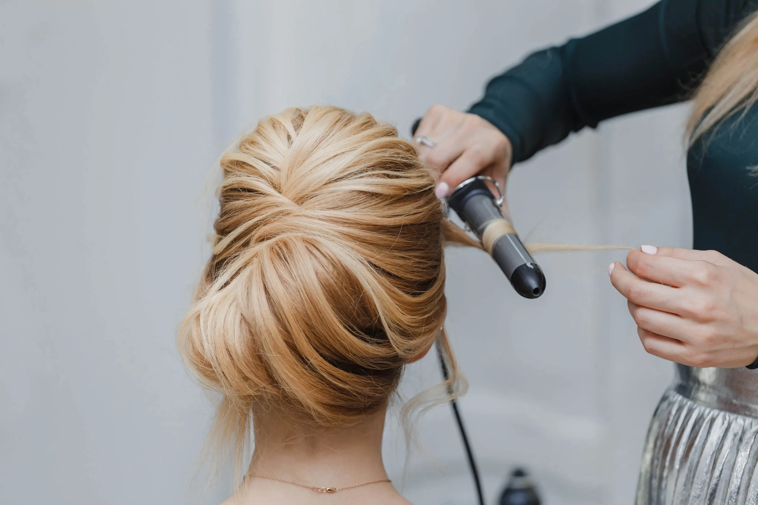 A person styles a woman's blonde hair in an elegant updo, using a curling iron to shape a strand at the back of her head.