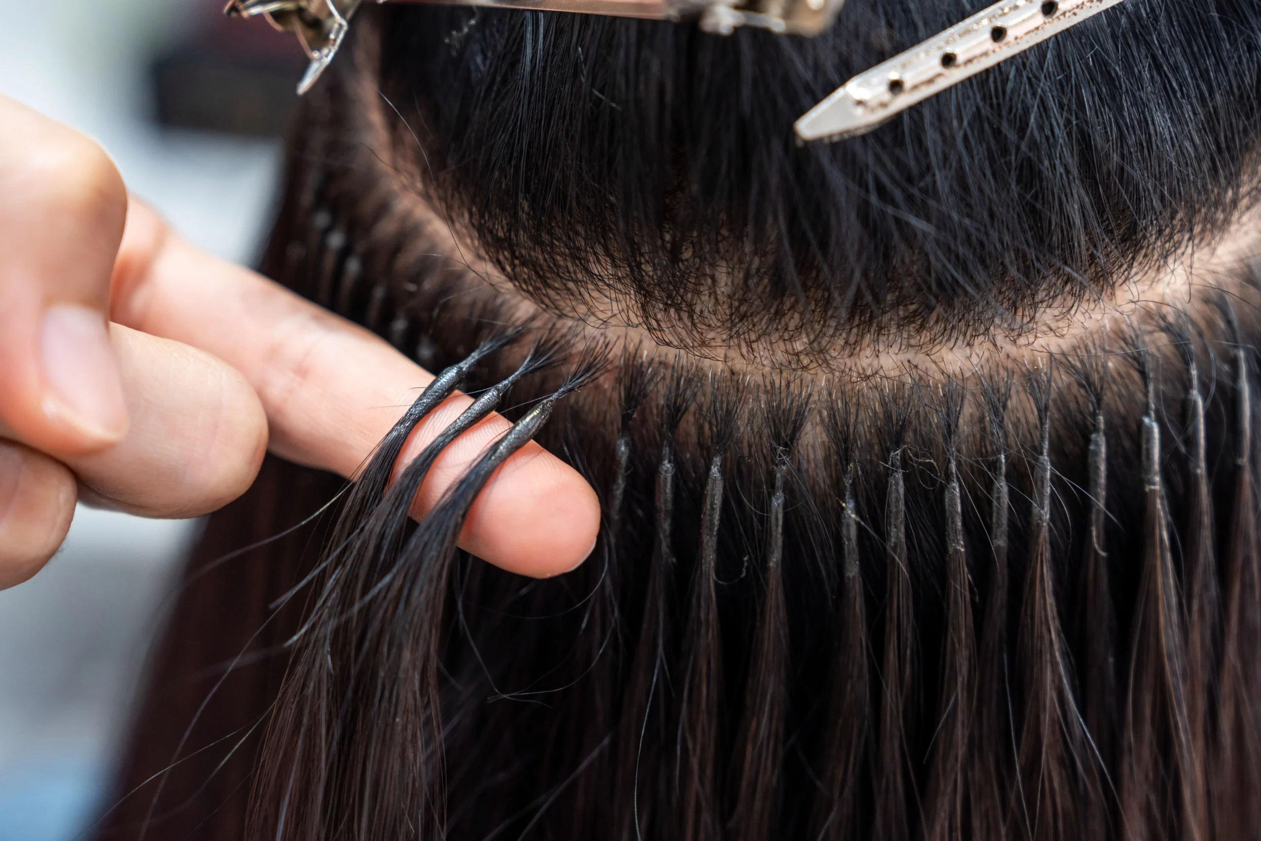 Close-up of a person getting hair extensions applied; strands of hair are attached near the scalp using small metal rings and clips.