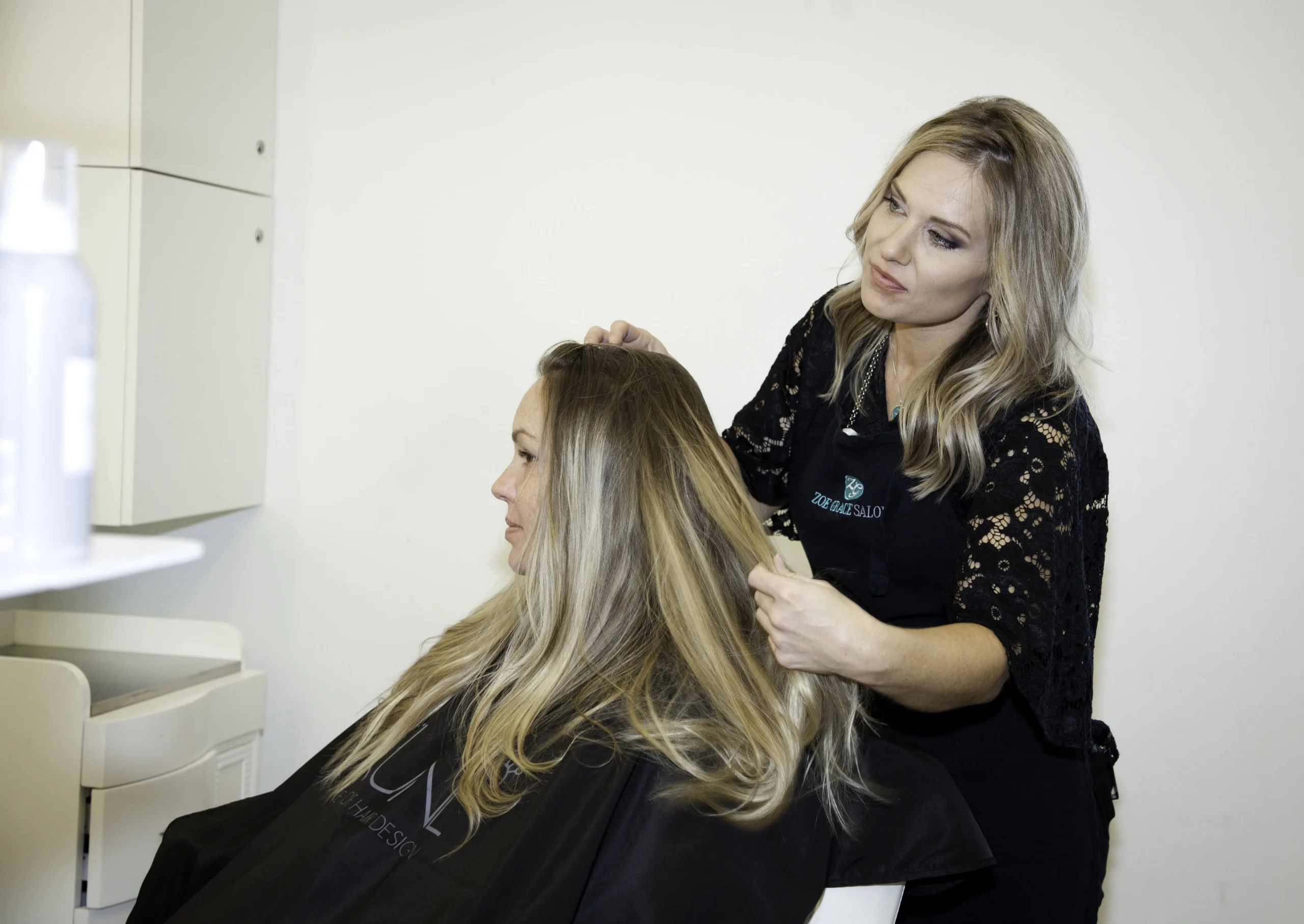 A hairstylist examines the hair of a seated client in a salon, both women have long blonde hair and are wearing black capes or aprons.