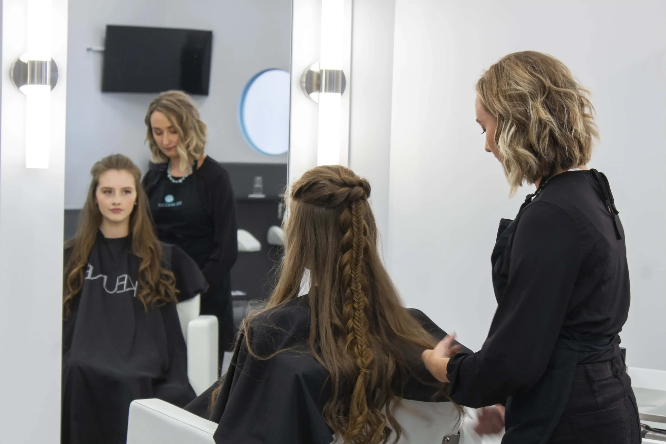 A hairstylist stands behind a seated woman with long hair, styling her hair in a salon with white walls and large mirrors.