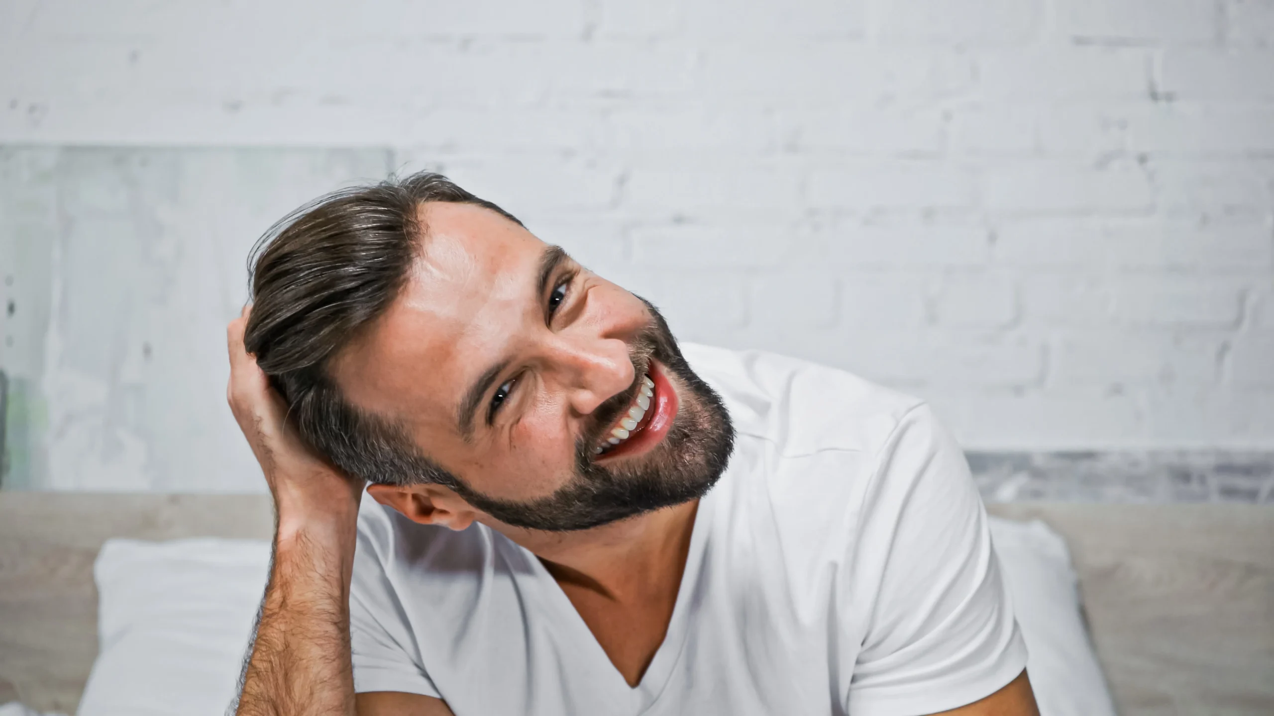 Man with short brown hair and a beard smiles while sitting on a bed, wearing a white t-shirt and touching his head with one hand. White brick wall in the background.