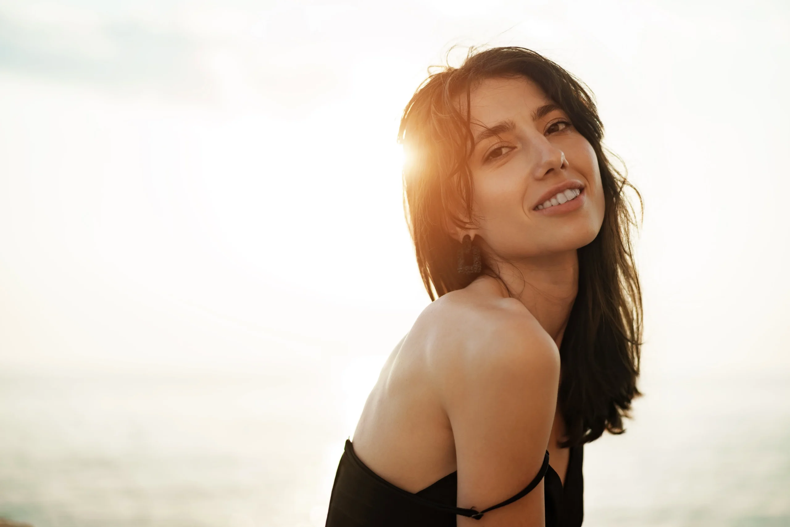 Woman with long dark hair in a black top smiles at the camera outdoors with sunlight shining behind her near a body of water.