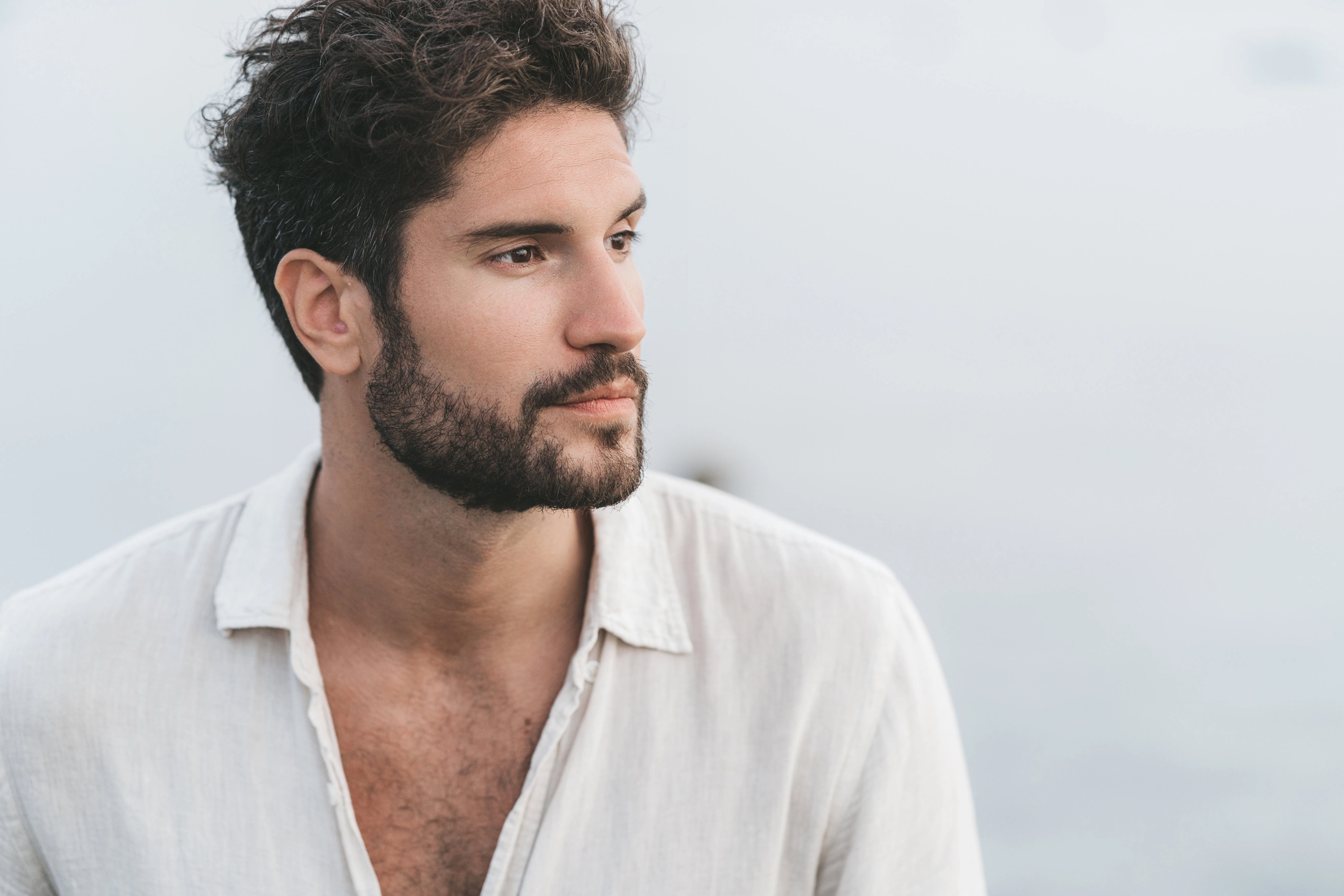 Man with dark curly hair and beard wearing a light-colored linen shirt looks thoughtfully to the side against a soft, blurred outdoor background.