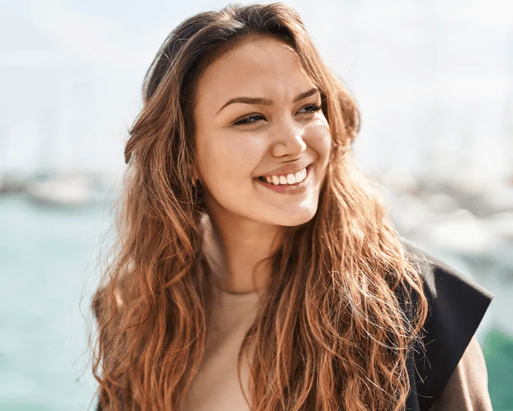 A woman with long wavy hair smiles while standing outdoors near a marina with blurred boats in the background.